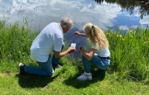Alexander Hess und Karin Böpple tropfen etwas Weihwasser aus Lourdes in den Moorweiher. Dafür knien beide im grünen Graß am Ufer des Sees.